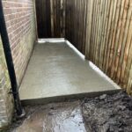 Freshly poured wet concrete in a narrow garden pathway between wooden and brick walls, with visible mud and puddles nearby.
