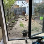 View from an open door showing a messy garden with scattered tools, patches of soil, and artificial grass near a fence at the back.