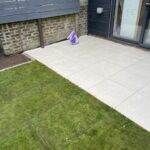 A tidy patio with large beige tiles next to artificial grass, featuring a purple shopping bag near the house door.