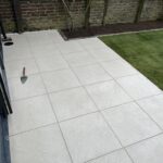 Newly installed light gray patio tiles with a trowel on them, adjacent to a garden bed and artificial grass lawn, bordered by a brick wall.