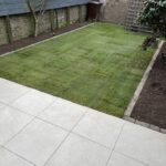 A neatly trimmed artificial grass lawn bordered by tiled patio, a flowering bush on the right, and a garden shed in the back.