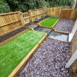 A newly designed garden with wooden planters, synthetic grass sections, grey paving stones, and purple pebbles on a brick patio.