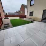 A modern backyard with large white tiles and a small artificial grass area, flanked by residential buildings on a cloudy day is transformed into an elegant space featuring a brick patio with integrated sections of artificial grass,