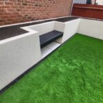 A small outdoor courtyard with a built-in bench, artificial grass, and an adjacent soil bed, bordered by white walls.