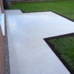 Newly laid large white patio tiles next to a lush green garden with a gravel border along a brick house.