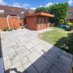 A well-maintained backyard featuring a large patio, a wooden shed, artificial grass, and a sunny sky.