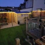 A nighttime view of a small garden with artificial grass and wooden fencing, illuminated by warm string lights.